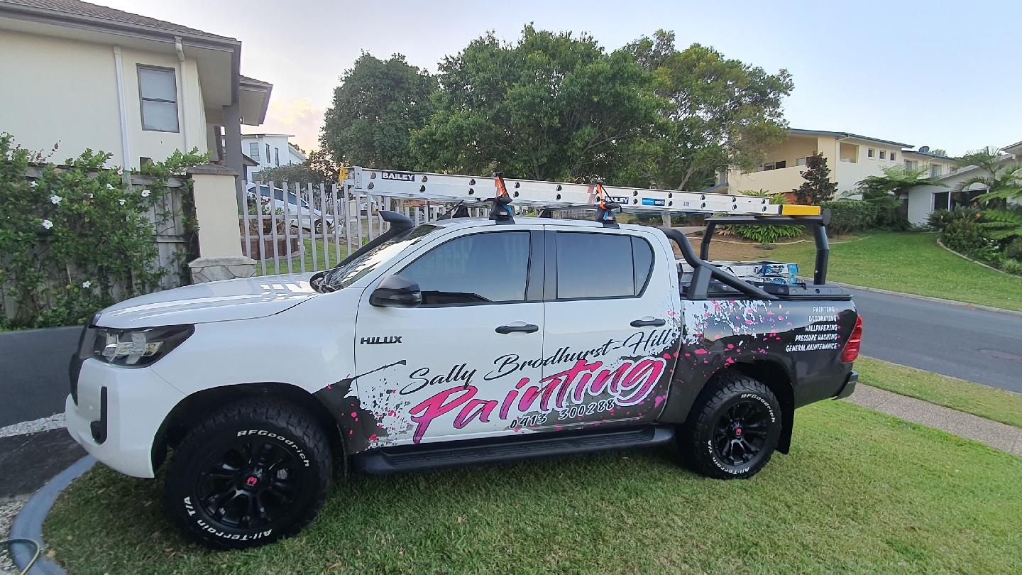 Sally's white HiLux ute with ladders and pink signwriting, parked on the kerb